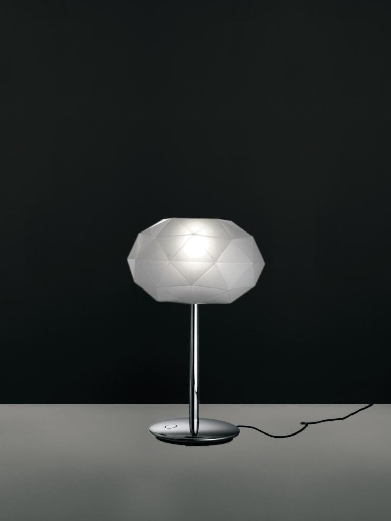 The Soffione table lamps from Artemide provide diffused halogen lighting. The diffuser, designed to recall the geometric structure of crystals, is made of white, handblown glass with a polished silk finish. The base is die-cast aluminum with a steel stem and is available in polished black or polished chrome. A dimmer switch is located on the cord. Two versions of the lamp are available: the first measures 22-3/16" tall and 9-1/16" wide and takes a 150W bulb; the second is 28-3/4" tall and 10-3/4" wide and takes a 250W bulb. Soffione was designed by Michele De Lucchi. artemide.us
