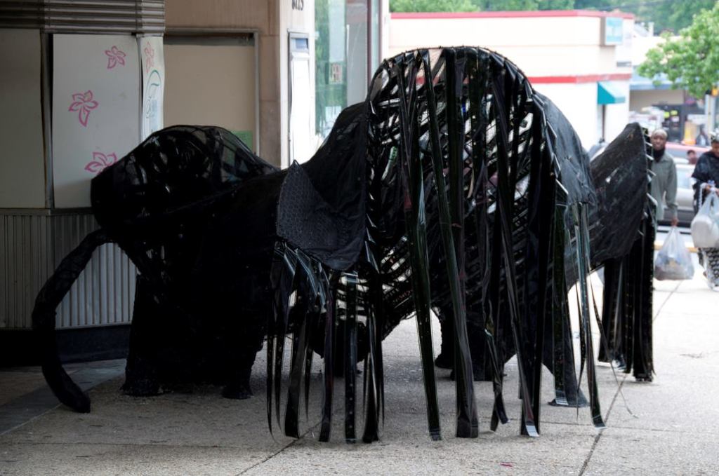 Movie Cave  Located in front of the long-defunct Flower Theater, which itself is the subject of an ongoing series of charrettes aiming for revitalization, the Movie Cave is a dark, parametrically designed sculpture covered in strips of film. The materials look opaque from without, but once you crawl inside, the backlit strips are revealed to be actual film from a reel containing one of the films in the Twilight series. The experience is completed by an auditory component: a looped track of movie theme songs playing from a hidden speaker inside the boarded-up ticket booth. “It looks like a wood mushroom,” Grenier says, noting that he enjoys the fact that “this is a dead theater and now there’s something growing out of it.”