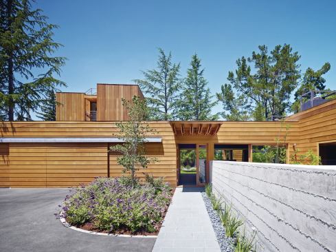 The landscaped entrance to the wood-clad Low/Rise House is at the end of a Vermont slate walkway, and flanked by a board-formed concrete wall that separates parking from private garden; the wall’s formwork was re-used as floor joists.