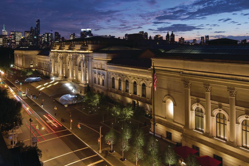 A nighttime view of the Metropolitan Museum of Art façade and plaza with the New York City skyline in the background.