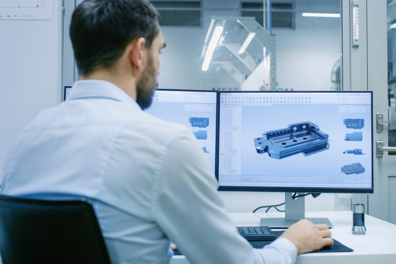 Engineer/ Technician Working on a Personal Computer with Two Displays, He's Designing New Component in CAD Program. Out of the Office Window Components Manufacturing Factory is Seen.