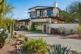 Crafting a harmonious blend of contrast and allure, the two-story addition to this home captivates. White exterior walls adorned with adobe brick accents pay homage to the original design, elegantly contrasting with black-framed windows, glass double entry doors, roof tiles, and gate to the outdoor haven. A wrap-around balcony above and front landscape areas graced by agave, cacti, and palms evoke natural charm. The exposed wood beams beneath the balcony and firewood storage infuse rustic elegance into the scene.