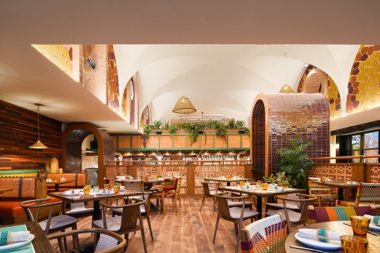 Tile archways lead to a raised dining platform with custom, hand-painted wall tiles. Yellow, cream, orange, and purple acoustic panels clad the walls, reminiscent of kernels of heritage corn, serving to incorporate core ingredients of the cuisine seamlessly into the visual language of the space. 