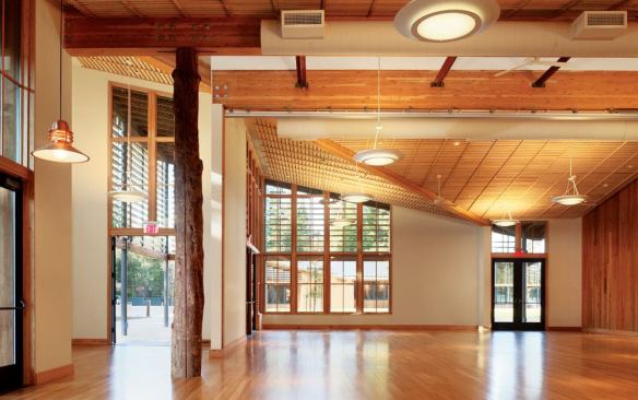 The community room in the town center's community hall is a multipurpose space designed to allow for meetings, parties, and other events. Supporting the roof is a tree column—an unusual construction that wraps a steel-pipe support with the split trunk of one of the trees felled to make way for the complex's sports fields. Multiple doors and windows can be opened to make the room an indoor/outdoor space during the milder months.