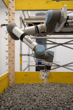 A robotic arm positions the granular, rock-like material around layers of textile filament.