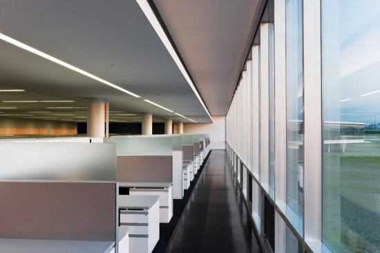 The open office space in the single-story portion of the building has cubicles with translucent partitions to allow daylight from the floor-to-ceiling windows to penetrate into the space. Circulation paths hug the glazed perimeter walls, with their operable windows to promote air circulation. Lighting and HVAC needs are centralized in an Armstrong TechZone ceiling system over the cubicles.