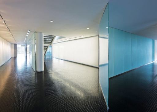 Running the length of the building is a frosted glass wall that serves as the barrier between the public and private zones of the building. Since much of the information being discussed in the office areas is proprietary, the glass serves as a means of obscuring visual and aural information without cutting off daylight. Here, the wall begins to separate an open office space (at right) from a small showroom (at left). The staircase leads to the second floor cafeteria and outdoor roof deck.