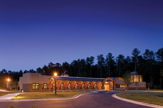 The indirect exterior lighting reflects the warm glow of the natural materials while articulating the architectural composition: the entry, the circular library (seen to the far right), and the peaked glass roof of the interior meditation room (seen in the background behind the colonnade).