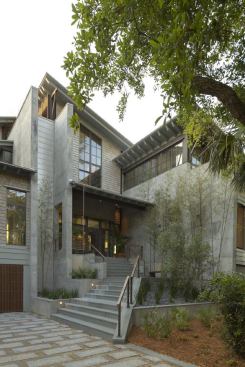 Concrete walls flank the front entry, blending in with the soft gray of bleached cedar shingles. Metal panels clad a slim elevator shaft.