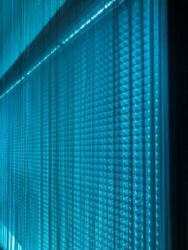 The team worked collaboratively to create a three-dimensional lighting effect for the walls while resolving acoustical needs for the space. The resulting juxtaposition of the textured acoustical back wall and the coil drape front metal scrim create a compelling moir?? effect that causes the walls to subtly shimmer as you move through the space. This effect can be made more dynamic through theatrical manipulation of the intensity and color of the light, or more static by turning the LEDs off completely, allowing the ambient light in the room to softly front light the scrim. 