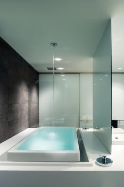 The infinity tub is filled from the ceiling and overflows into the lit space around it. A porcelain tile floor folds up the back wall.