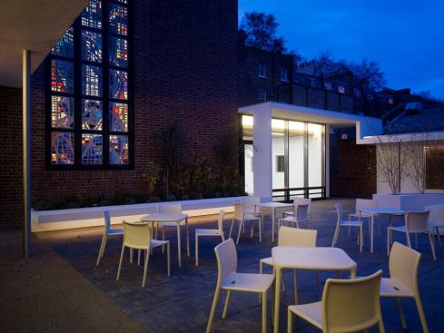 The stained glass window and East Community room beyond as seen from the garden. The community rooms use direct linear fluorescent and linear fluorescent cove lighting. The indirect lighting of the cove creates an architectural connection between the community rooms and the garden. The use of indirect lighting continues in the garden under the benches and in the bench alcoves providing subtle lighting to the garden seating. In the summertime, the community rooms can be opened into the garden creating one continuous space.