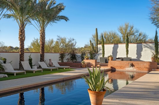 Graceful palms sway, casting dappled shadows upon the glistening pool. Chaise lounges invite relaxation on the soft expanse of green grass. At the far corner, a newly integrated spa harmoniously merges with the surroundings, appearing as if it has always belonged.