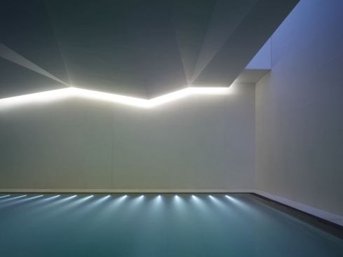 A midnight-blue line of light and the pool's ladder handrail are allowed to reflect perfectly in the water below by turning off the pool LED lighting. Daylight from the skylight is reflected back across the angular ceiling above, rendering the surfaces and displaying the ceiling's dynamic sculptural form.