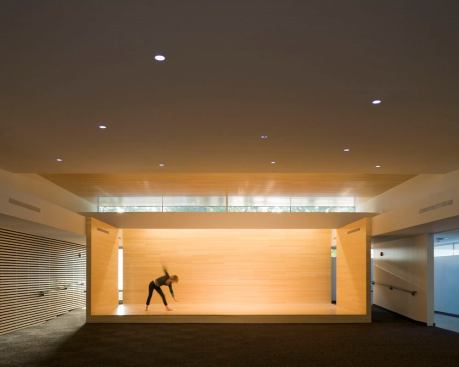 The stage is illuminated with a combination of PAR38 tracklights concealed above the ceiling, linear fluorescent coves that wash the back stage wall, and indirect illumination of the wood ceiling above the stage.