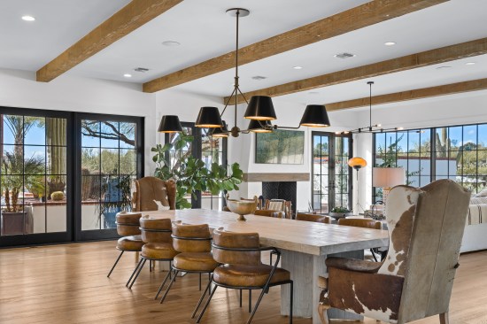 Around the substantial rectangular dining table, cowhide motif wingback chairs grace the table's ends, while eight caramel-colored leather cushioned chairs offer comfort. The living space showcases an inviting fireplace. Black-framed double doors flank the hearth on both sides, leading to the outdoor yard. Peering through the slider reveals the forthcoming outdoor oasis: palms, a saguaro, and an array of cacti thriving in raised planters.