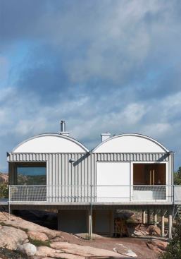 The roof’s twin barrel vaults echo the rounded forms of the topography. Siding is white-painted cement board and corrugated cement board panels; the roof, corrugated metal.
