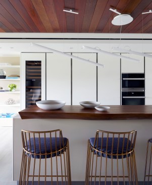 Thin polished brass stools with navy slate blue cushions from Haute Living adorn the near side of the island. Above the island, a purposely angled white LED linear pendant from Vibia provides an additional architectural layer to the space.