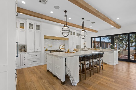 The kitchen dazzles in white, highlighted by gold, black and wood accents. From the pot filler to the wood beam hood and plumbing fixtures, the details shine. A farmhouse sink and floor-to-ceiling cabinets complete the scene. An 8-burner range top adds a grand touch. 