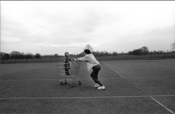 Grandmother pushing grandchild through a parking lot in Clapham Common, South London, 1996.