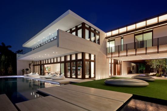 Seen from the rear, the home features laminated mahogany windows and door frames, water elements, and a glass-fronted second level. The metal brise-soleil, canopies, and louvered walls feature a powder-coated paint in a woodgrain texture. 
