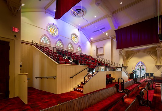 View of new tiered balcony seating and restored stained glass windows. The original wood pews were restored, and new cushions were provided on the balconies and as movable seating at the lower level. The hanging draperies provided needed absorptive materials to address acoustical concerns. Slot diffusers provide conditioned air along the perimeter of the space, while the original oculus locations in the ceiling serve as return air registers. Fresh air is provided through louvers in the gable of the new elevator tower addition.