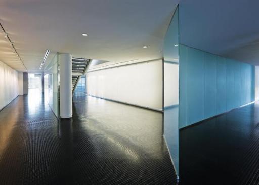 Running the length of the building is a frosted glass wall that serves as the barrier between the public and private zones of the building. Since much of the information being discussed in the office areas is proprietary, the glass serves as a means of obscuring visual and aural information without cutting off daylight. Here, the wall begins to separate an open office space (at right) from a small showroom (at left). The staircase leads to the second floor cafeteria and outdoor roof deck.