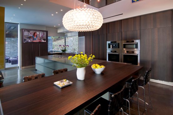 Modern kitchen design with dining room table of dark wood, cabinets and flush ovens, and TV wall.