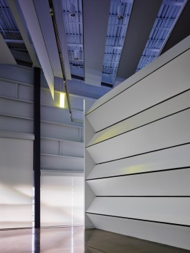 The translucent shades create soft lighting and ghostly shadows along the perimeter edge.  The undulating sound trap wall allows the space to be “tuned.”  Blue fluorescent lights add a romantic quality to the space.  One gold “note” is visible. “Notes” appear, move, change and disappear during the day capturing the kinetic energy within the space.
