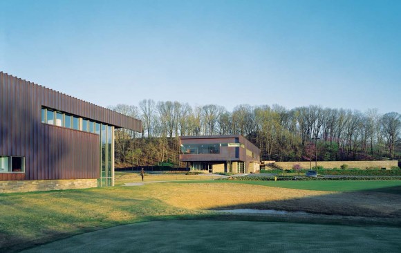 The structures surrounding the clubhouse employ the same material palette of glass, local stone, and copper. A copper-clad practice facility for the University of Arkansas golf team faces the clubhouse across a green. Stone-faced golf cart storage runs perpendicular to the clubhouse and stands only one story tall.