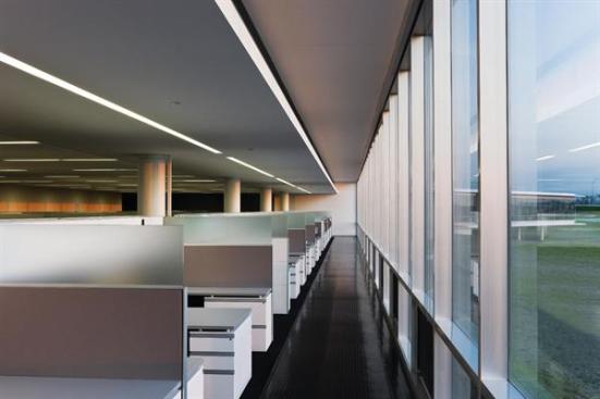 The open office space in the single-story portion of the building has cubicles with translucent partitions to allow daylight from the floor-to-ceiling windows to penetrate into the space. Circulation paths hug the glazed perimeter walls, with their operable windows to promote air circulation. Lighting and HVAC needs are centralized in an Armstrong TechZone ceiling system over the cubicles.