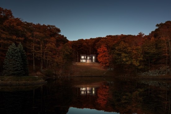 The home is illuminated at night, with the soft glow reflecting magically on the pond’s surface.