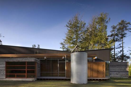 A high roof slopes down from each end to a trough that sends rainwater to a metal cistern. A flat lower roof is planted with native grasses.