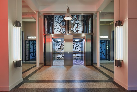 A view of the glass-walled elevator, which reveals a mural by typographic artist Pieter Ceizer on the back shaft wall. In the foreground, refurbished fluorescent fixtures find new life as wall sconces.