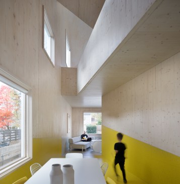 The double-height dining area on the first floor of Haus Gable features a bright yellow floor and walls that stand out from the exposed CLT-panel surface of the upper walls and ceilings above. The threshold of the master bedroom beyond is marked by a shift to gray accents.