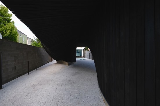 A cavelike carport under the J-House is lined with permeable pavers and gives access to both the main entry stair (at right) and back stair volume (at left); the renovated guest house can been seen past the pool beyond. 