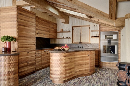 Caption: Kitchen interior with walls made using rammed earth, hemp, and straw, featuring carved walnut cabinets designed by Chaletbau Matti and Francis Kéré, blinds by Aïssa Dione, and a stool by Madoda-Fani.