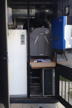 A look inside an exterior mechanical room, showing a heat pump (at left and lower right), a heat-recovery ventilator (top right), and a solar photovoltaic inverter (at right on the door).