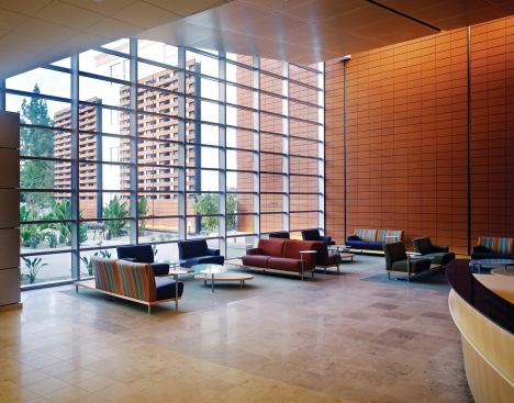 The main lobby has a floor-to-ceiling curtain wall system that admits ample natural light into the reception and waiting areas. A Moeding terra cotta façade continues into the building, where it is lit by wall washer light fixtures hidden above the canted artificial wood acoustical ceiling. Acoustical Wood Ceiling: Armstrong; Woodworks perforated metal ceiling panel in White Maple with a clear finish; armstrong.com - Floor Tile: Oregon Tile & Marble; Jura Stone honed in beige and gray; oregontileandmarble.com