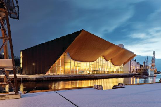 The geometric, dark-gray aluminum panels cladding the building’s rear and side elevations juxtapose the warmth and curves of the Kilden Performing Arts Center’s main façade, which emulates a rippling stage curtain or the wooden hulls of ships pulling 

into harbor.