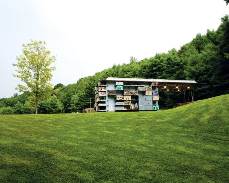 The barn's space-frame roof projects out over a paved area, providing a shelter for the storage of sand and other materials. An outdoor shower is a water source for wetting materials and a place for the owner's family to rinse off after taking a dip in the nearby lake.