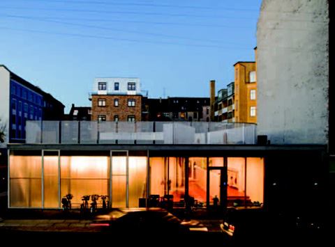 As dusk falls, lights in the building's southwest corner cast a glow behind frosted glass; children play on swings in this area during inclement weather. The entrance to the facility is to the right, and is indicated by a door in the third bay of the clear glass section.