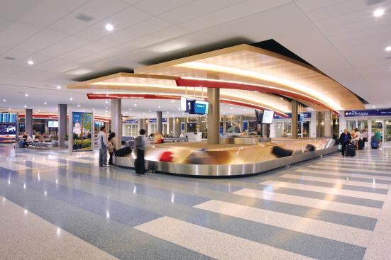 In the baggage claim area, a cove-mounted linear LED fixture uplights the wooden ceiling panels above each carousel.