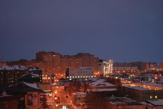 The illuminated Vershina Trade and Entertainment Centre stands out in the evening skyline of Surgut.