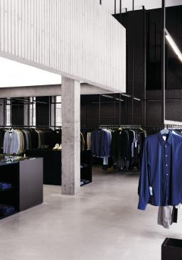 Clothes hang from a series of ceiling-mounted black steel frames in the double-height space around a corrugated-concrete mezzanine.