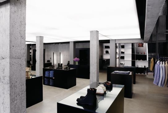 Underneath the mezzanine, shoes and other accessories are displayed on minimalist black pedestals under a backlit white opalescent stretch ceiling.