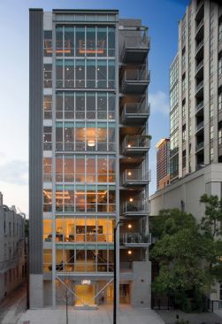 This concrete-and-steel building holds its own among taller neighbors, thanks to elegant cross-bracing, metal-screen walls, and commercial storefront windows. A lobby and parking area occupy the ground floor; eight levels of housing rise above.