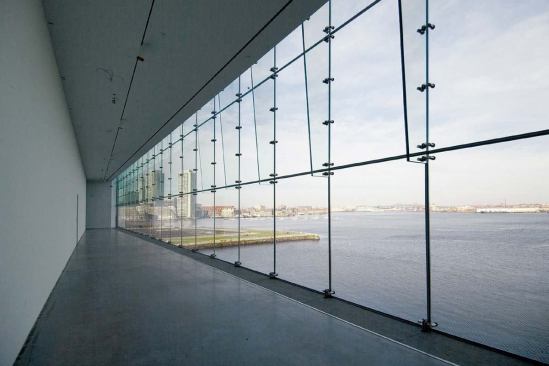 Founder's Gallery

The Founder's Gallery is a crossover space that spans the full width of the building's north face. By connecting the main east and west galleries, the linear room is woven into the choreography of the art-viewing sequence. The gallery's glass curtain wall-which stretches floor to ceiling, revealing a panoramic view of Logan Airport, Bunker Hill, and Boston's business district-is composed of cantilevered laminated glass fins with point support fittings. The wall was designed and tested in a full-scale mock-up to ensure its ability to withstand high winds whipping in from the Atlantic Ocean.