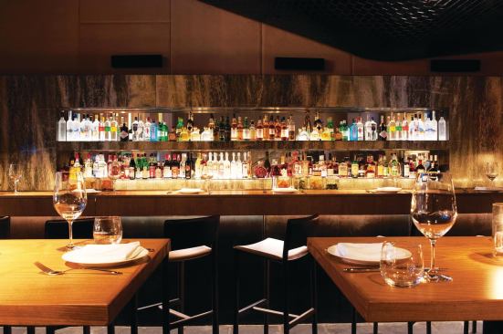 The lobby bar was designed to be a destination in and of itself. The bar proper is clad in stone panels and wood, with inset, mirrored, and uplit shelves for displaying the liquor bottles. The space connects to the restaurant, Faustina.