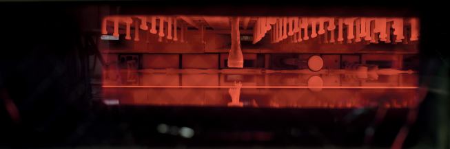 On the red-hot forming line, the glass is poured in a thin layer atop a bath of molten tin, from which it stays separate as it takes shape. T-shaped electrodes hang above to convey heat, and a guide wheel controls the stream's width, which in turn determines the glass's thickness.